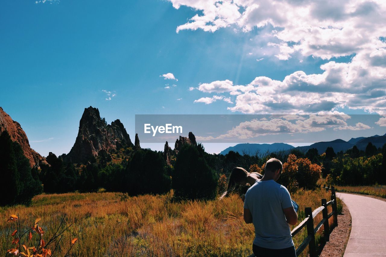 Rear view of man with woman by grassy field against sky on sunny day