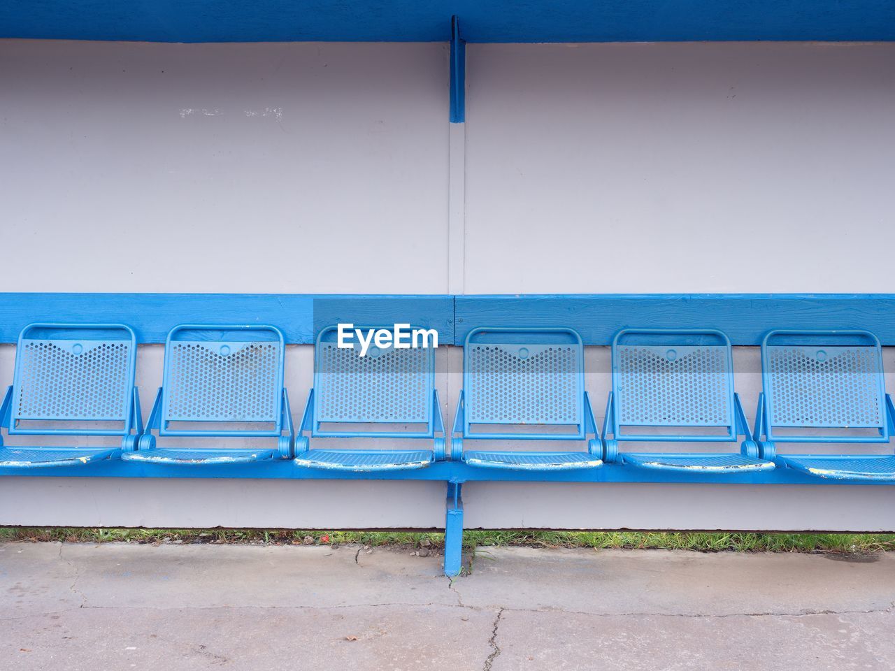 New blue metal seats on outdoor stadium players bench, chairs with blue paint below wooden roof.