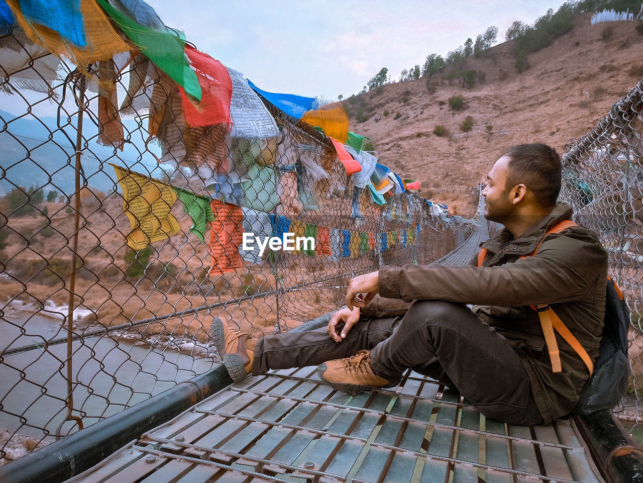 Side view of young man sitting on a suspention bridge with prayer flags outdoors