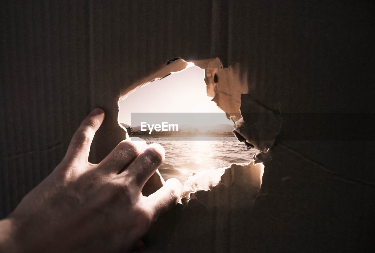 Close-up of hand tearing cardboard against sea during sunset