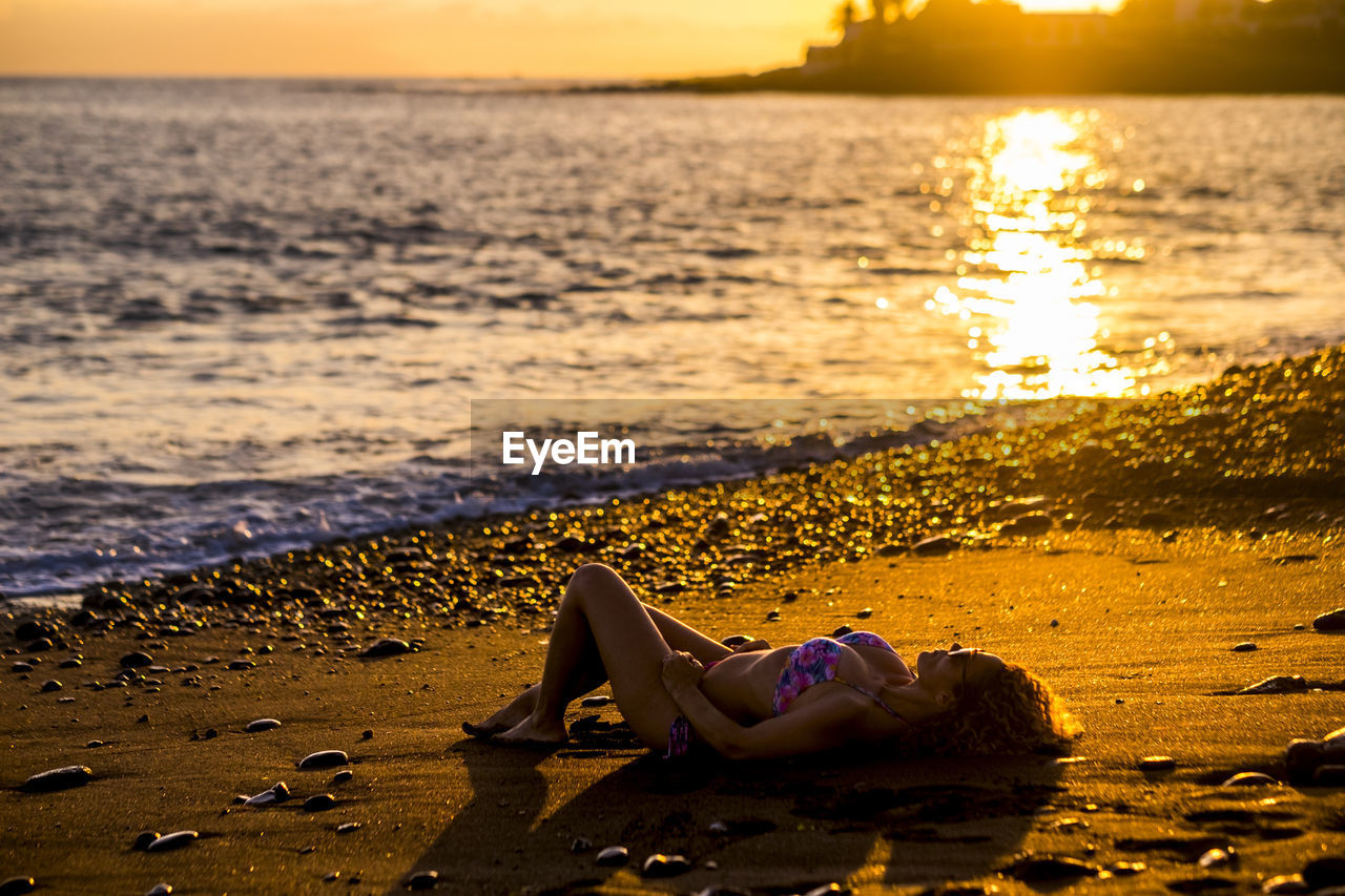 Full length of woman relaxing on shore at beach during sunset
