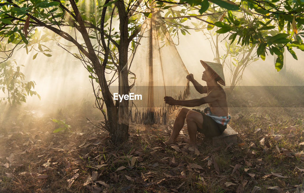 Man adjusting fishing net while sitting by tree