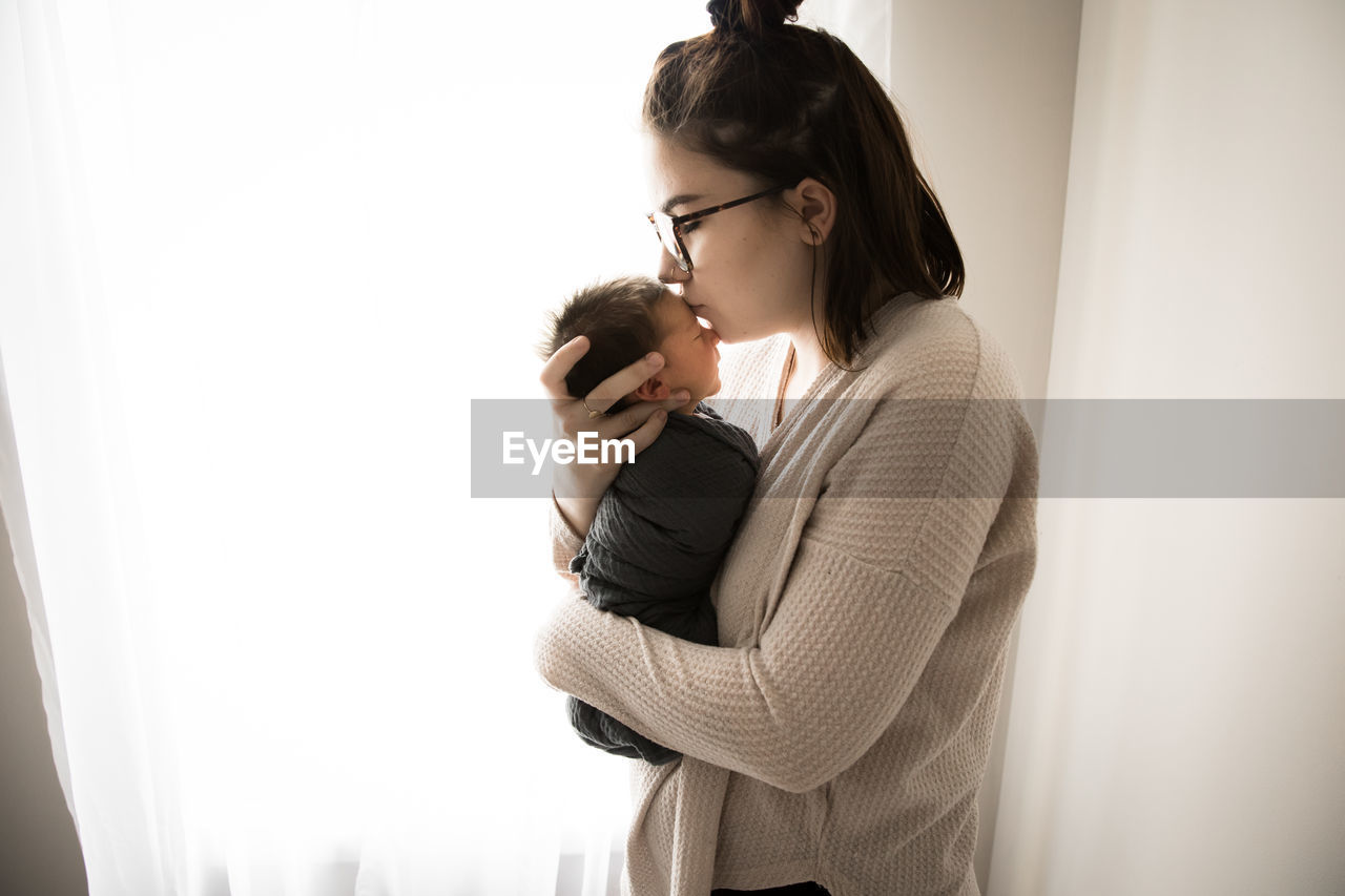 Backlit gen z hipster mom kisses newborn baby in front of window