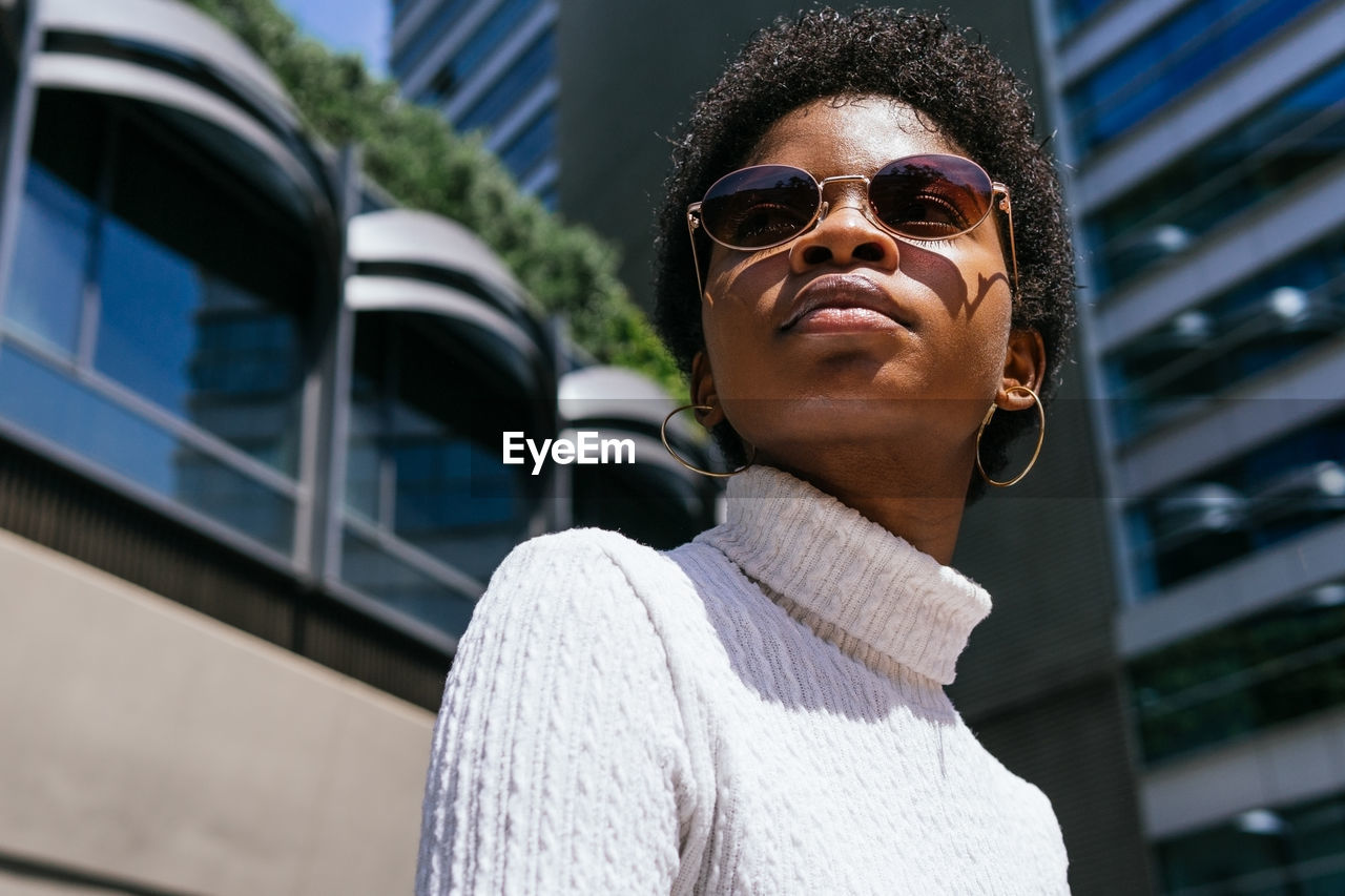 From below positive young african american woman in trendy outfit looking away and enjoying sunlight while sitting on bench on modern city street