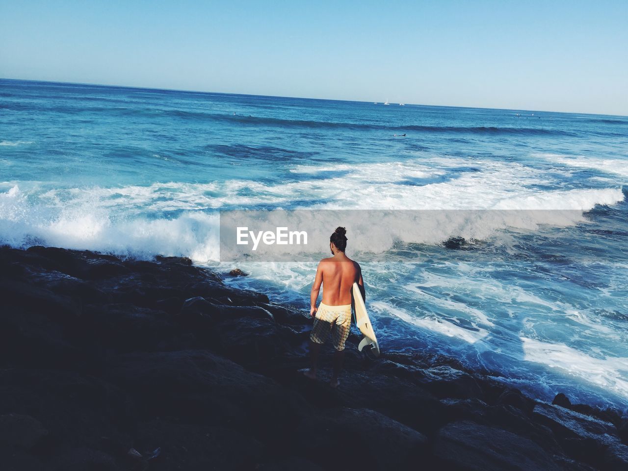 Rear view of shirtless man with surfboard standing on beach against clear blue sky