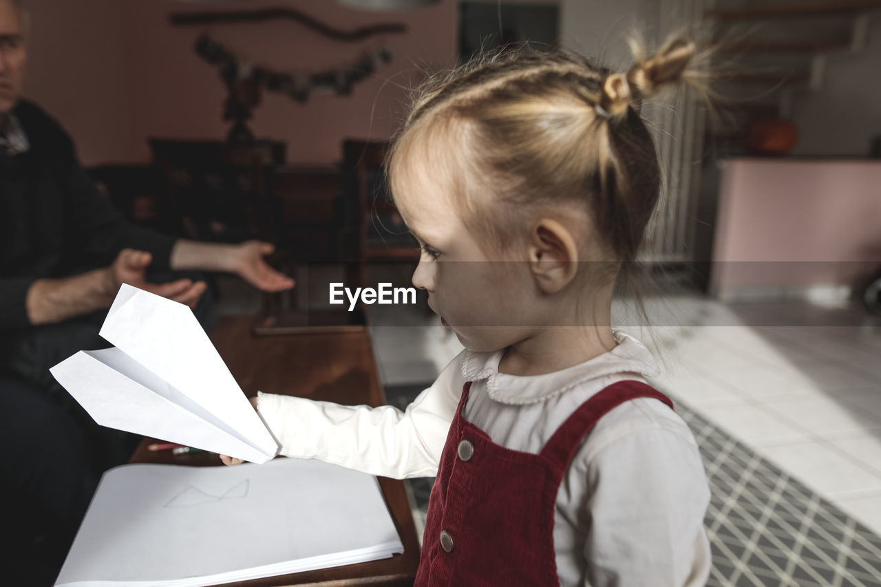 Girl holding paper plane in living room with grandfather in background