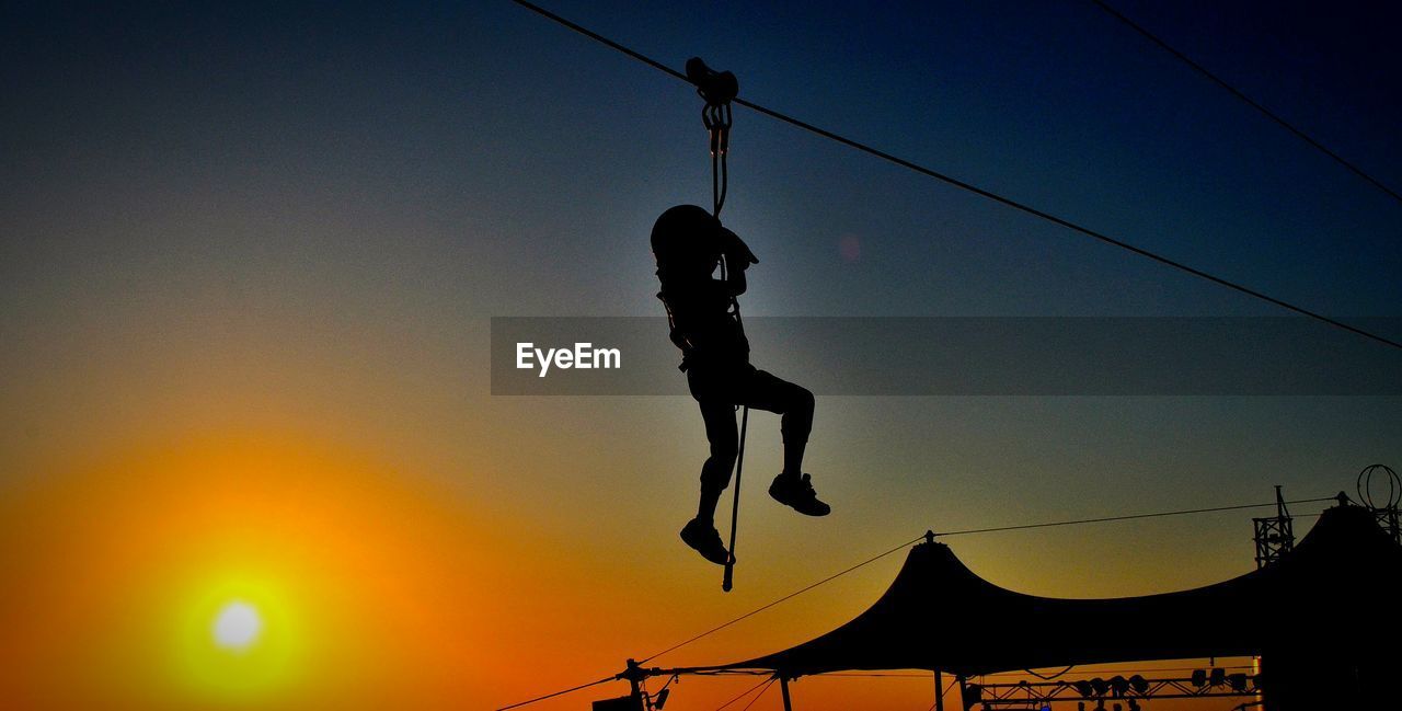 Low angle view of silhouette man zip lining against clear sky during sunset