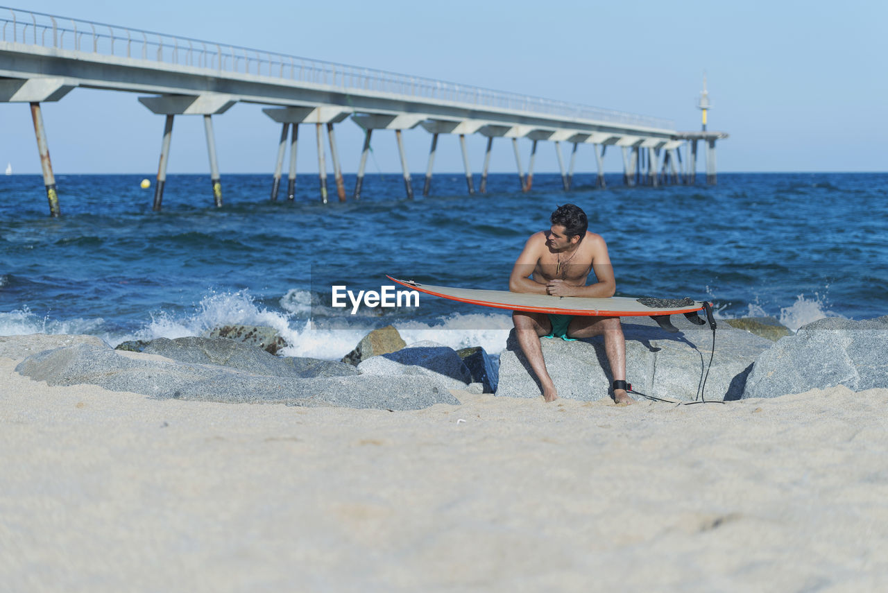 Shirtless man holding surfboard while sitting on rock at beach