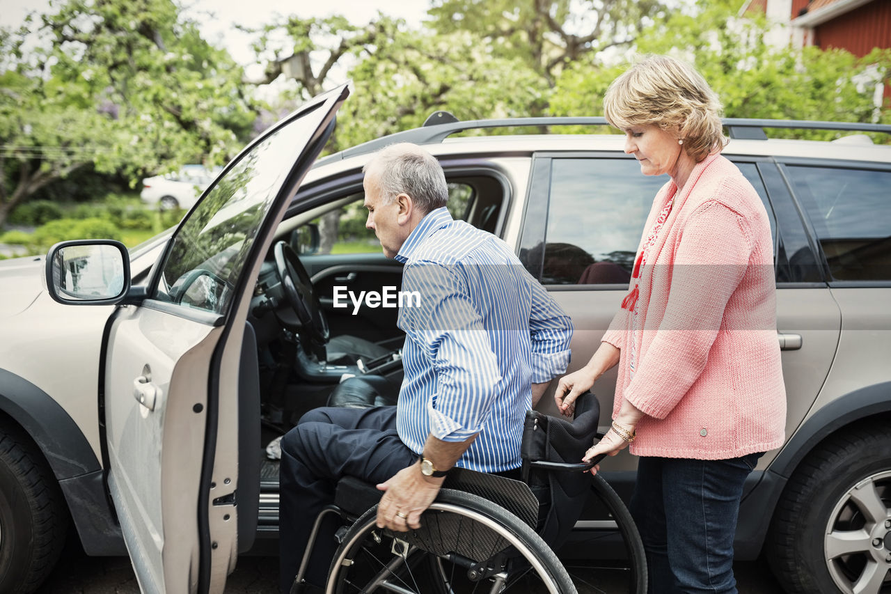 Disabled man sitting in wheelchair held by woman outside car