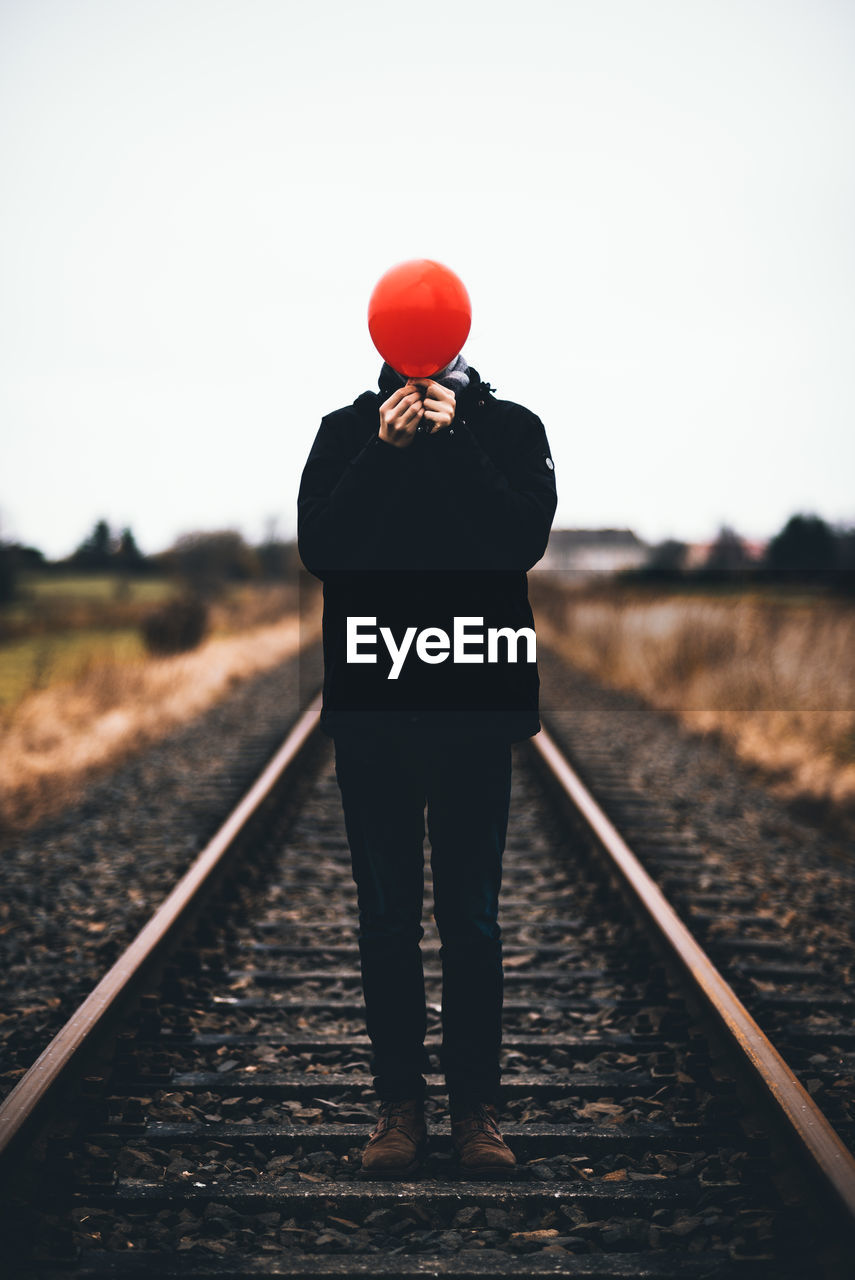 Man covering face with red balloon while standing on railroad track against sky