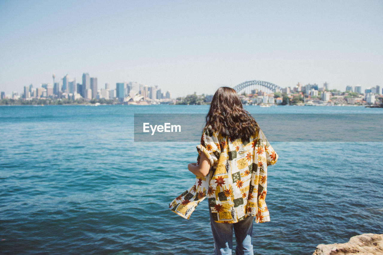 REAR VIEW OF WOMAN STANDING BY SEA AGAINST CITY