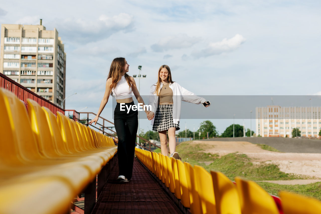 Two teenage girls walk together through the stands of the school stadium, talking, holding hands