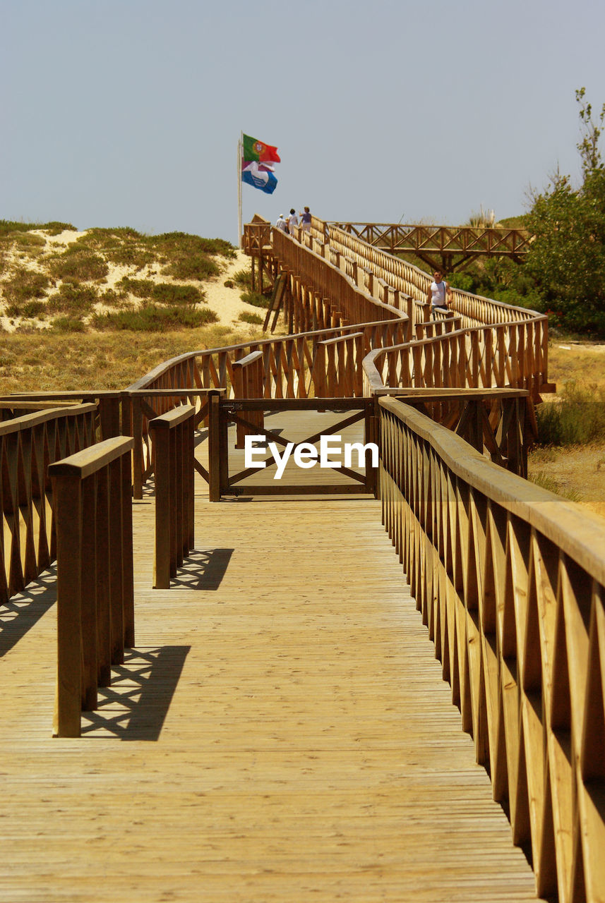 walkway, boardwalk, flag, sky, architecture, nature, patriotism, railing, wood, built structure, land, day, no people, clear sky, travel destinations, outdoors, vacation, pier, sunlight, sunny, bridge, water, beach, travel, footpath, scenics - nature, environment