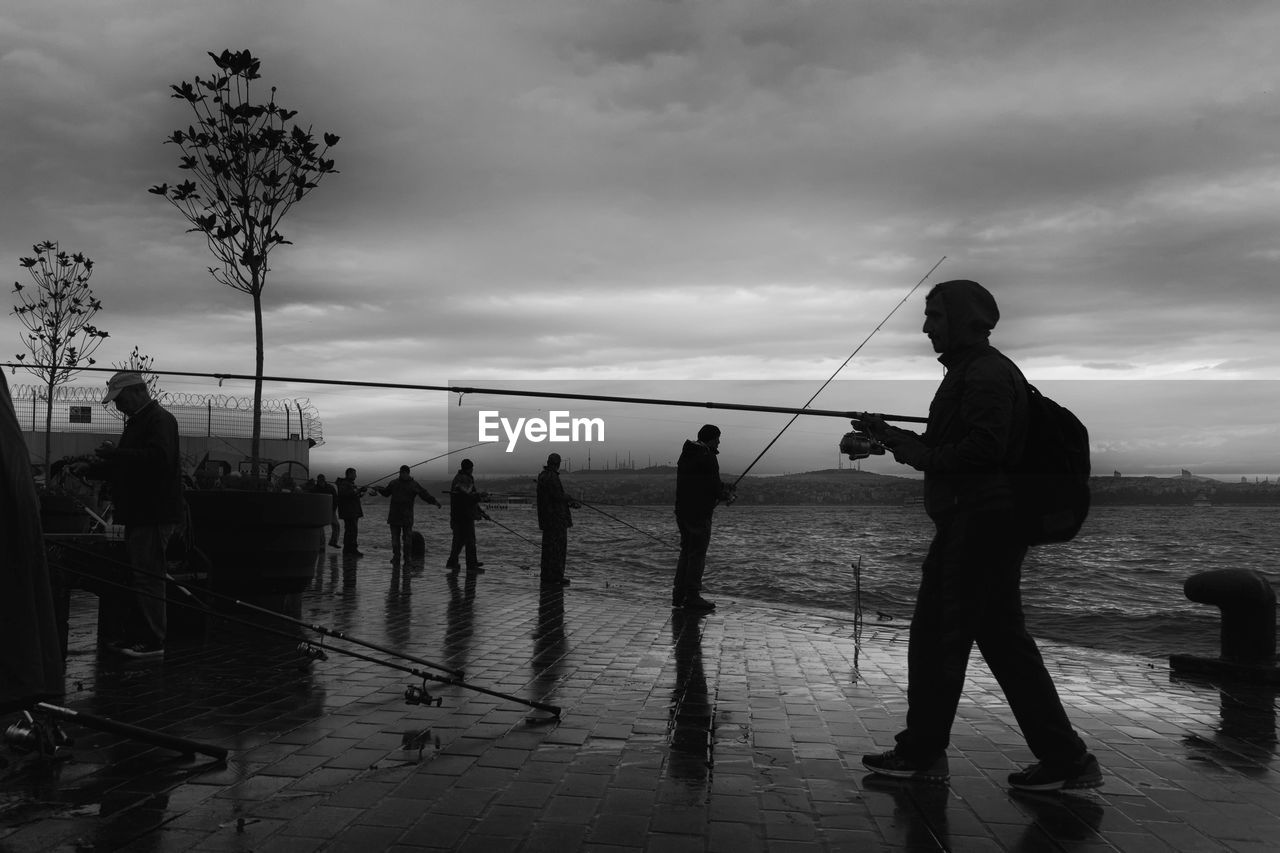 Silhouette people fishing in river on pier against cloudy sky