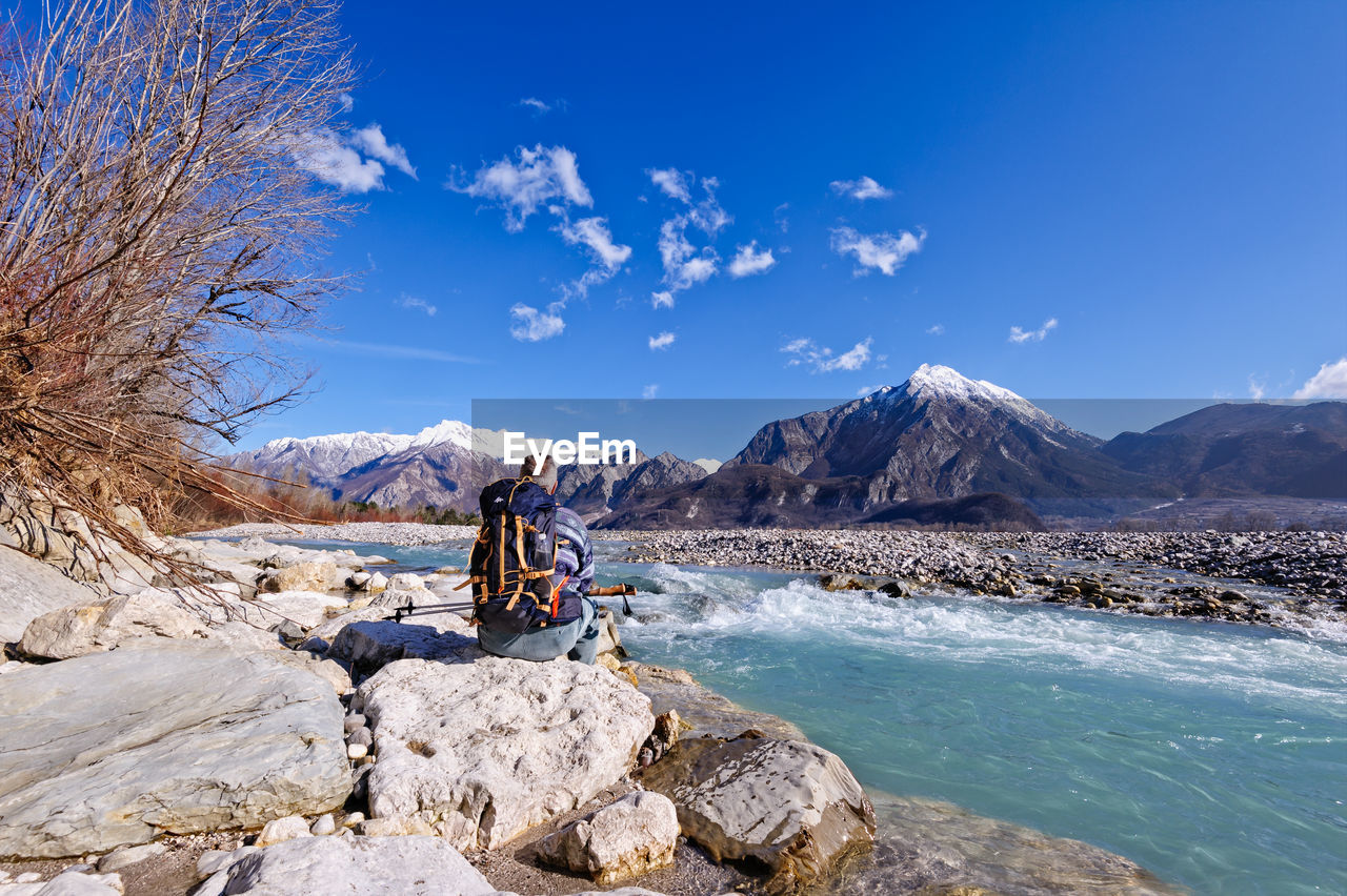 Rear view of man sitting rock by river blue sky