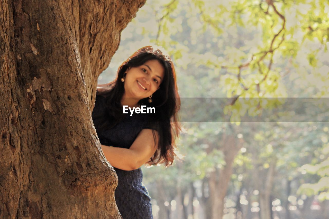 PORTRAIT OF SMILING YOUNG WOMAN STANDING BY TREE