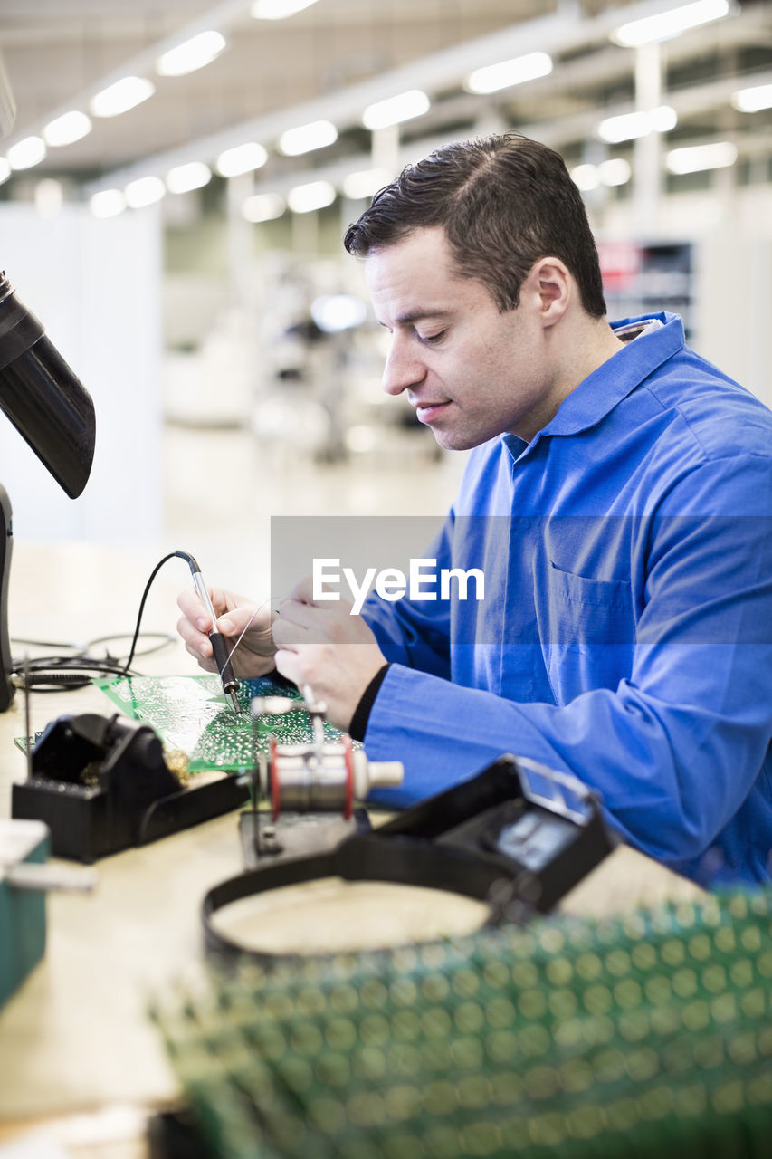 Mature male technician soldering circuit board at desk in industry