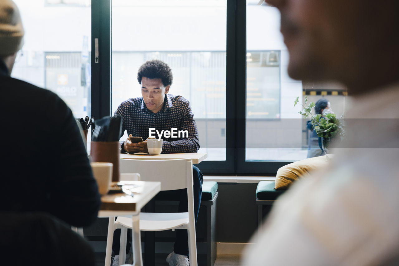 Young customer using mobile phone while sitting with coffee cup at table in cafe