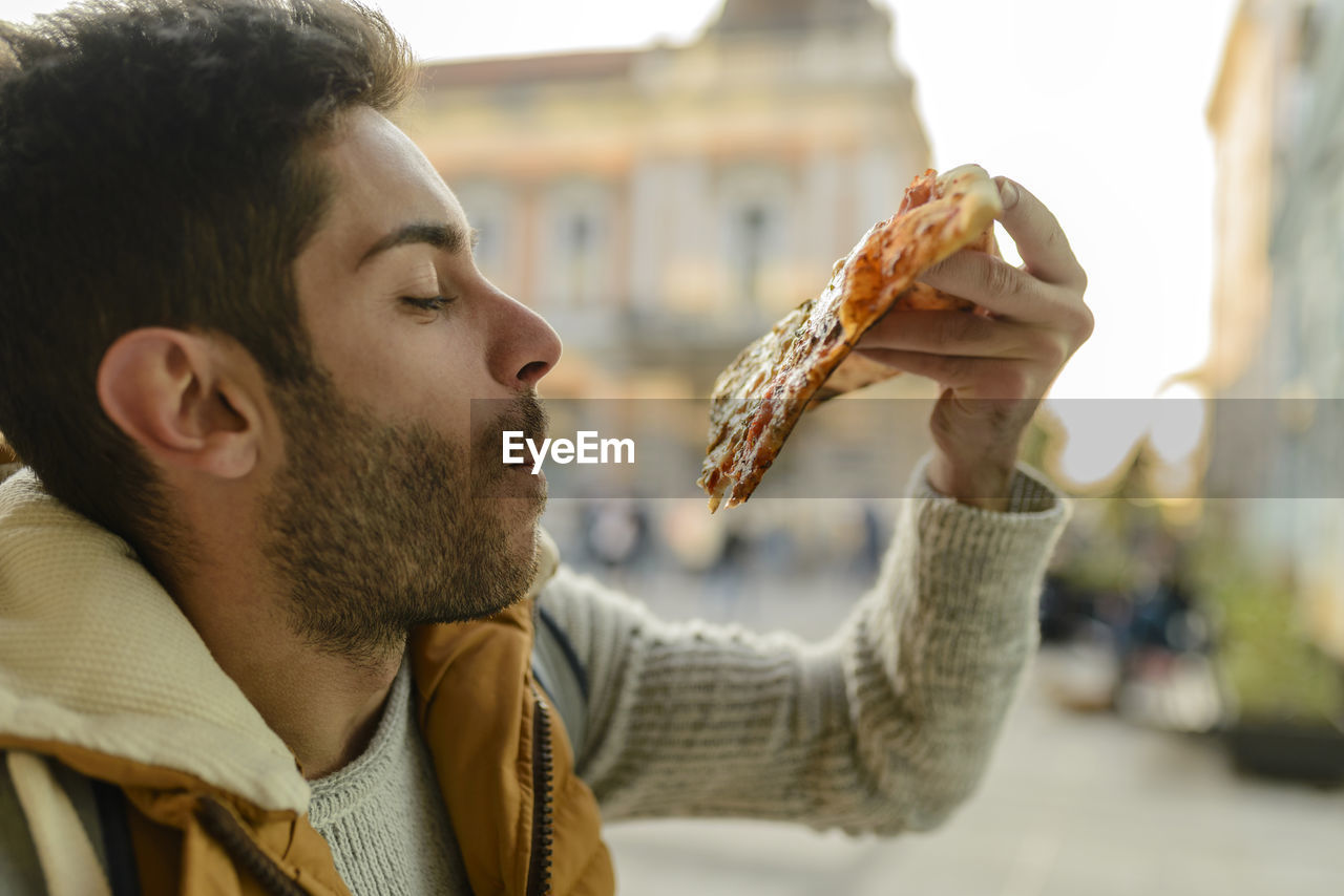 Close-up of young man eating pizza while standing in city during sunset