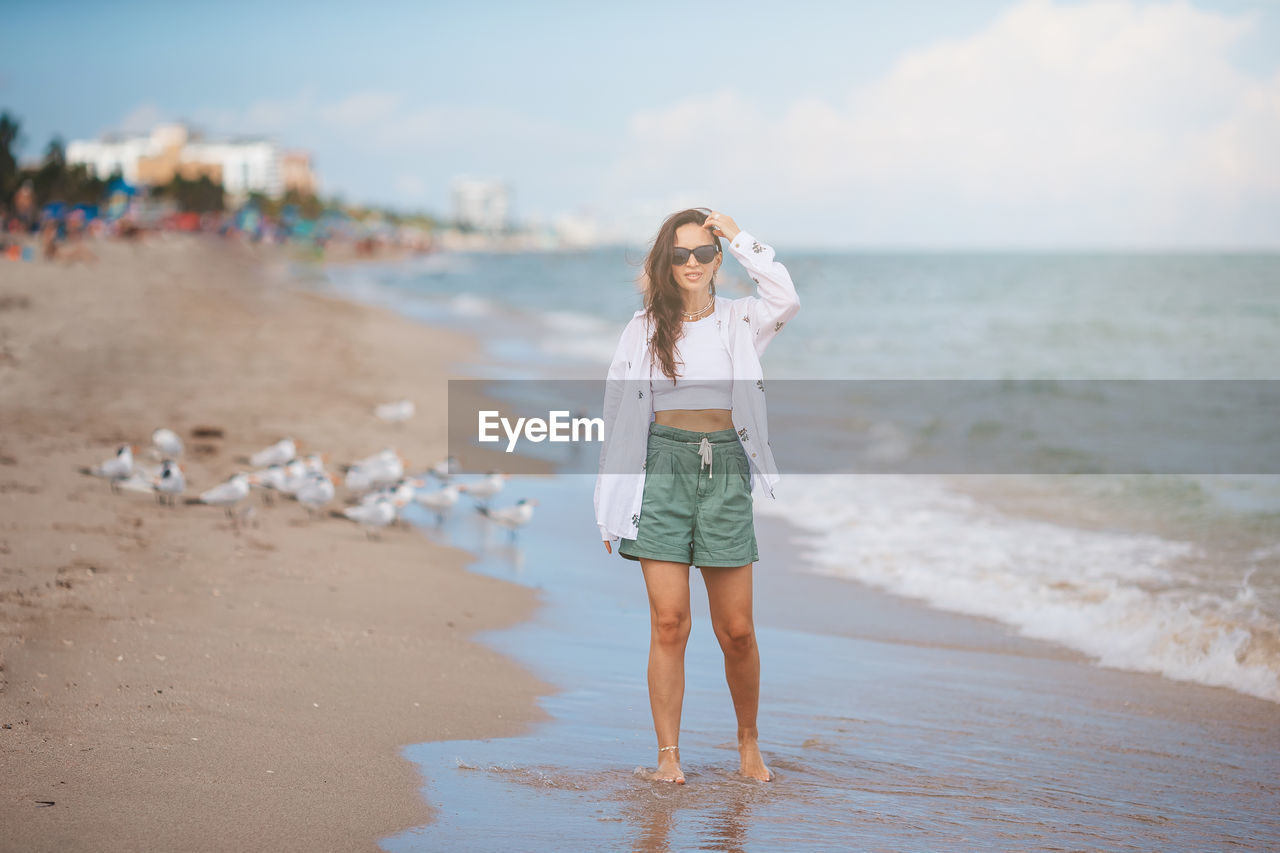 beach, land, water, sea, one person, women, sky, holiday, sand, nature, full length, vacation, trip, adult, long hair, hairstyle, clothing, body of water, young adult, summer, ocean, shore, casual clothing, happiness, emotion, leisure activity, fashion, standing, smiling, walking, front view, day, barefoot, blond hair, travel, travel destinations, portrait, lifestyles, blue, coast, outdoors, child, carefree, female, person, beauty in nature, motion, focus on foreground, cloud, enjoyment, childhood, tourism, dress, fun, spring, relaxation, cheerful, looking at camera, looking, positive emotion