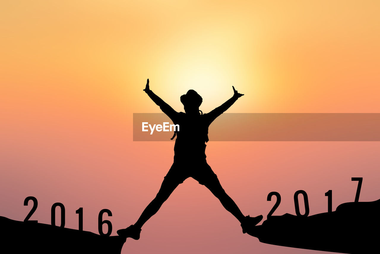 Silhouette person with arms raised standing on rocks with numbers against sky during sunset