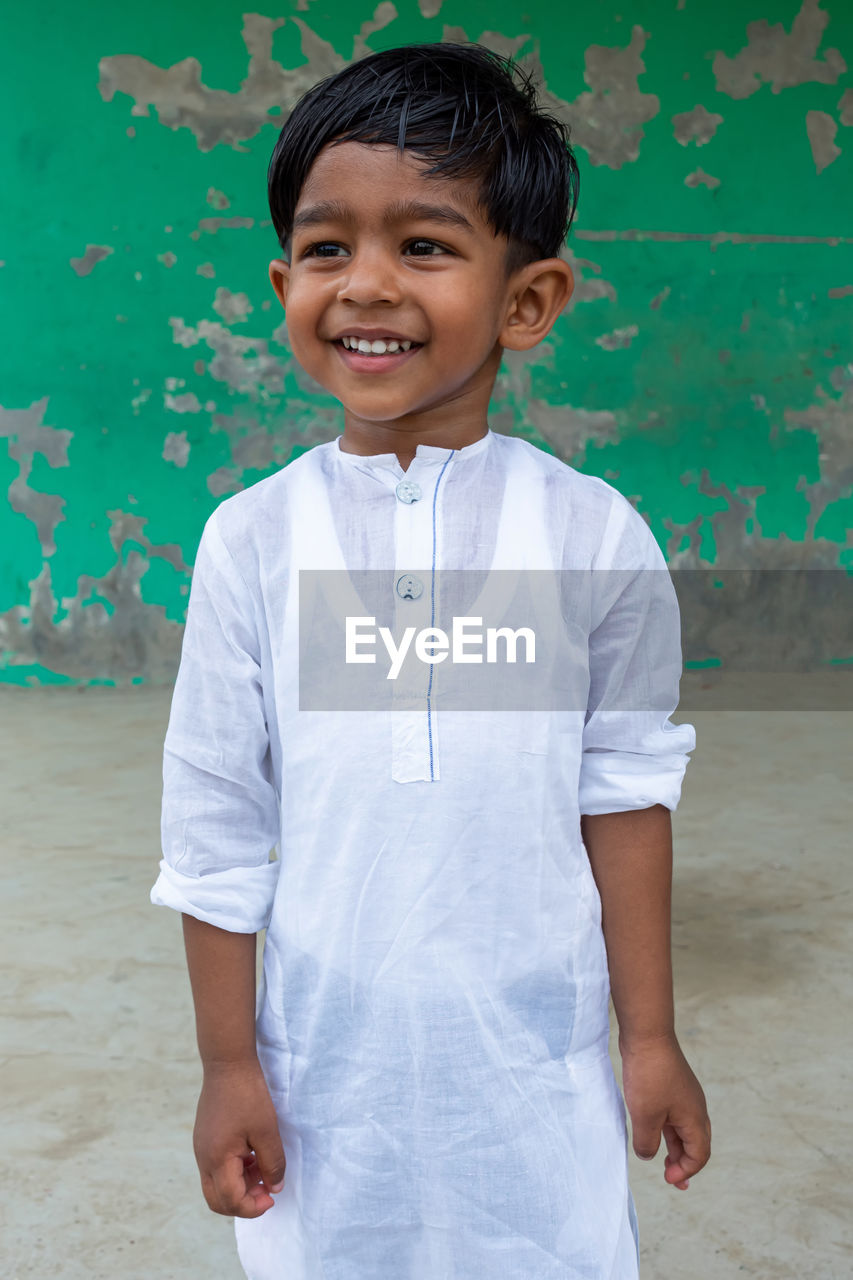 PORTRAIT OF SMILING BOY STANDING AGAINST WALL