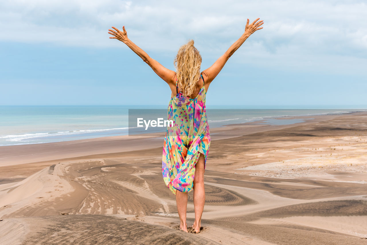 Back of an adult blonde woman standing on the desert gesturing freedom with the arms raise