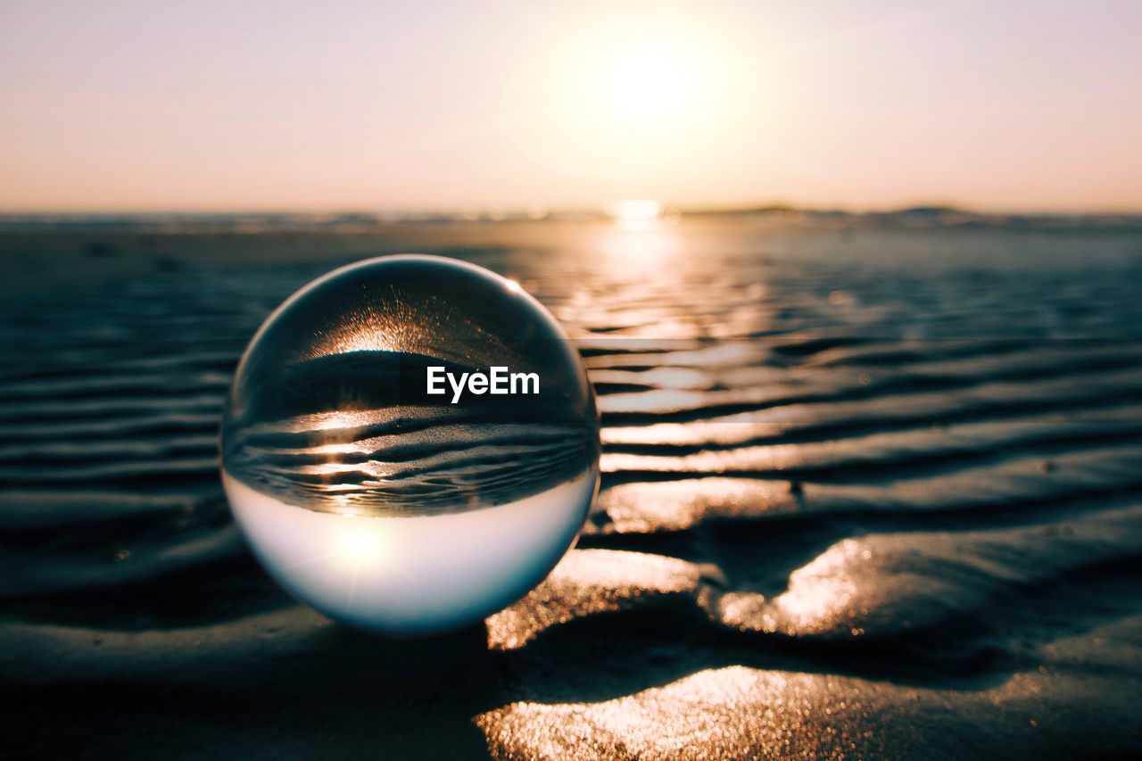 reflection, sky, sunset, water, sea, nature, sunlight, light, tranquility, sphere, land, beauty in nature, blue, close-up, beach, scenics - nature, macro photography, crystal ball, sun, tranquil scene, no people, focus on foreground, outdoors, horizon over water, horizon, idyllic, shiny, dusk, environment, clear sky, single object, transparent, relaxation, summer, holiday, copy space, shape, sand, shadow