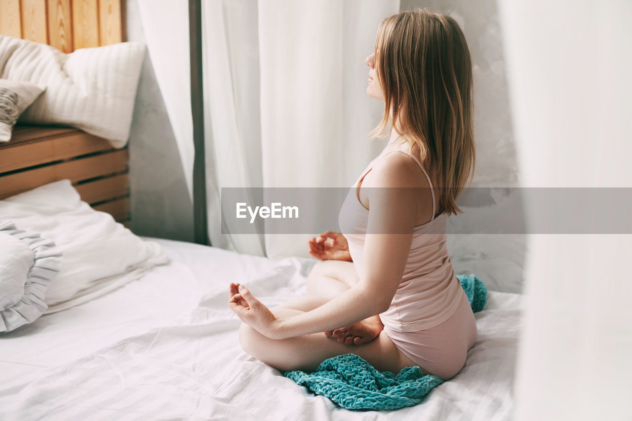 A young woman in pyjamas is sitting on a bed in a lotus position 