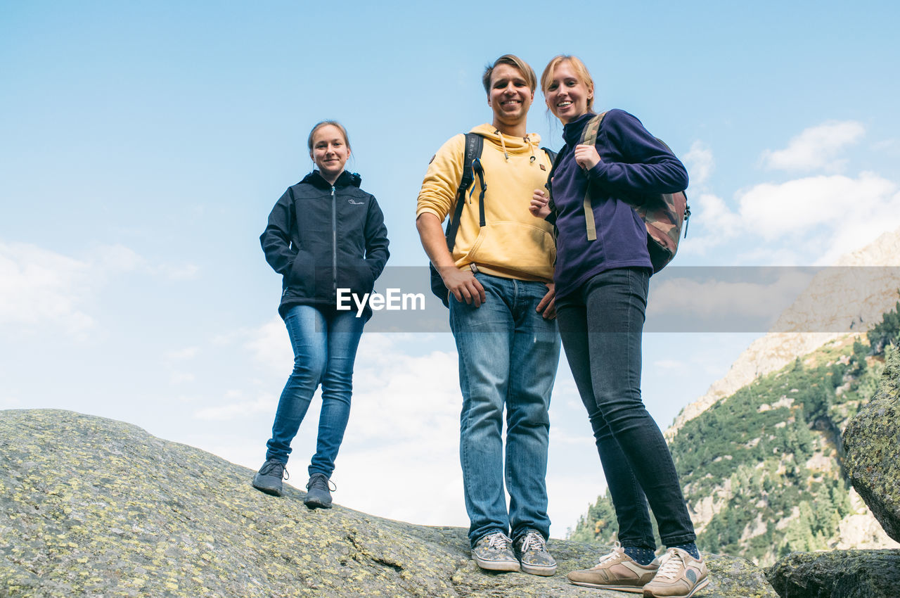 Full length portrait of friends standing on rock against sky