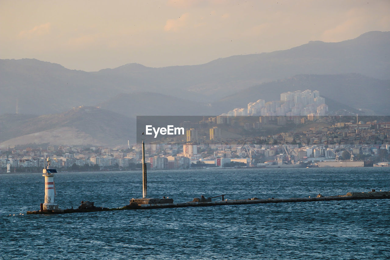 Lighthouse in sea by city and mountains against sky