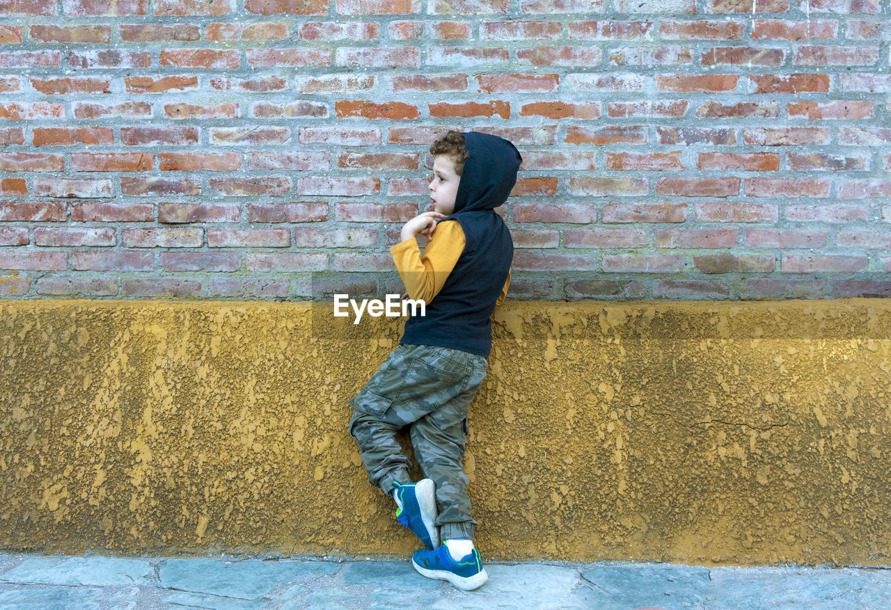 Child in hooded sweatshirt and camouflage pants leaning against brick wall