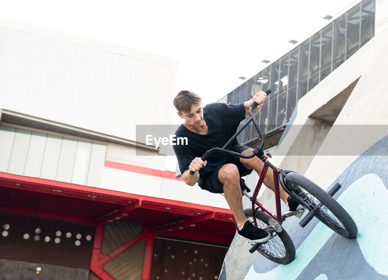 Young male in casual clothes riding concrete wall on bmx bicycle while spending time in skate park in weekend