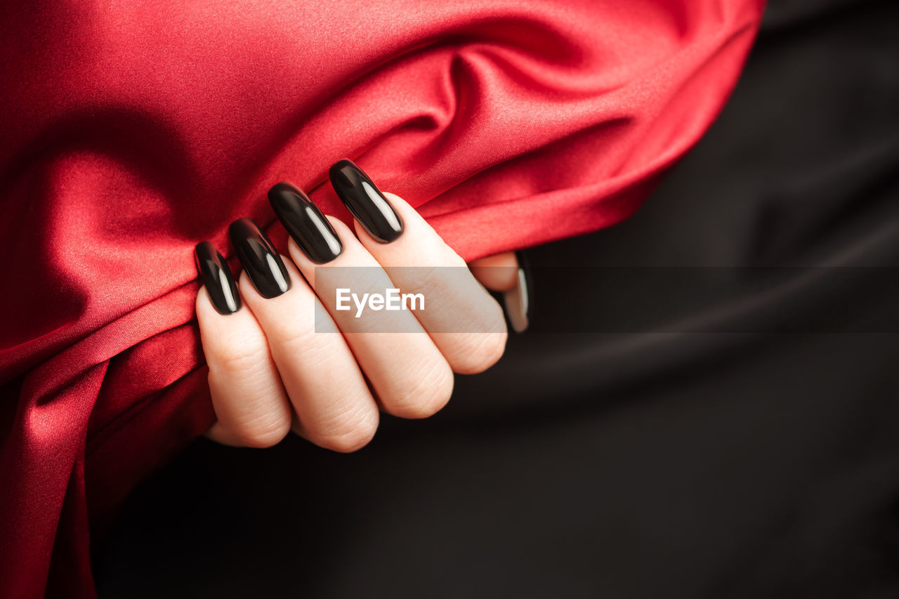 Hands of young girl with black manicure on nails holding red satin cloth on black background