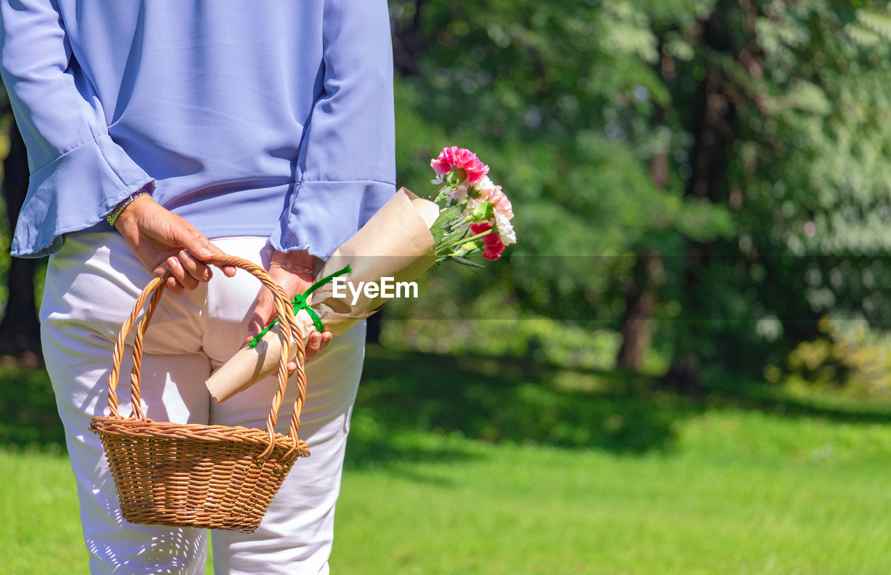 Midsection of woman holding wicker basket and bouquet while standing in park