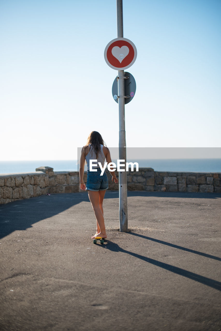 REAR VIEW OF WOMAN STANDING AT SEA SHORE AGAINST CLEAR SKY