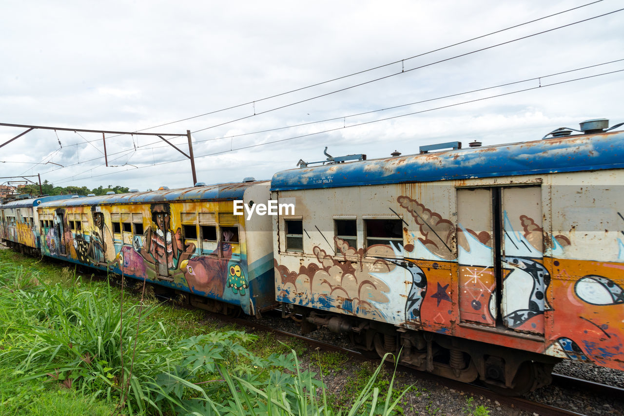 rail transportation, transportation, train, transport, mode of transportation, railroad track, track, vehicle, railway, locomotive, public transportation, sky, railroad car, cloud, rolling stock, land vehicle, nature, travel, day, architecture, grass, no people, cable, passenger car, graffiti, electricity, outdoors, passenger train