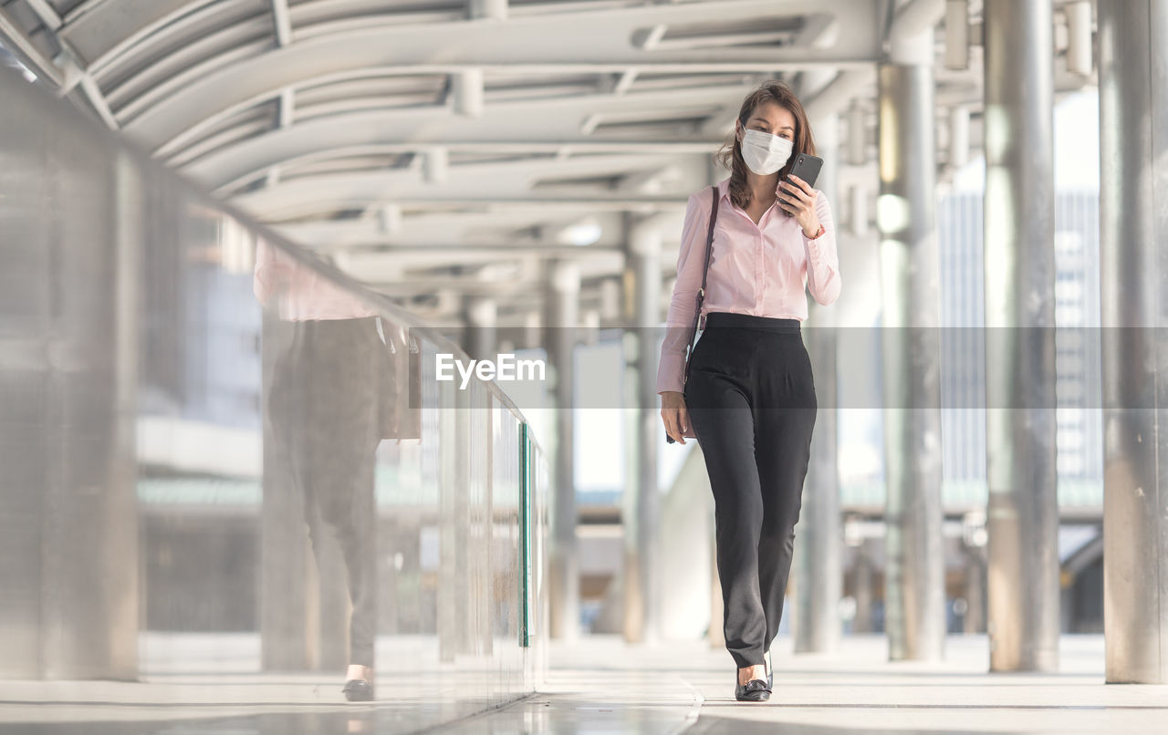 Full length of businesswoman wearing mask using mobile phone outdoors
