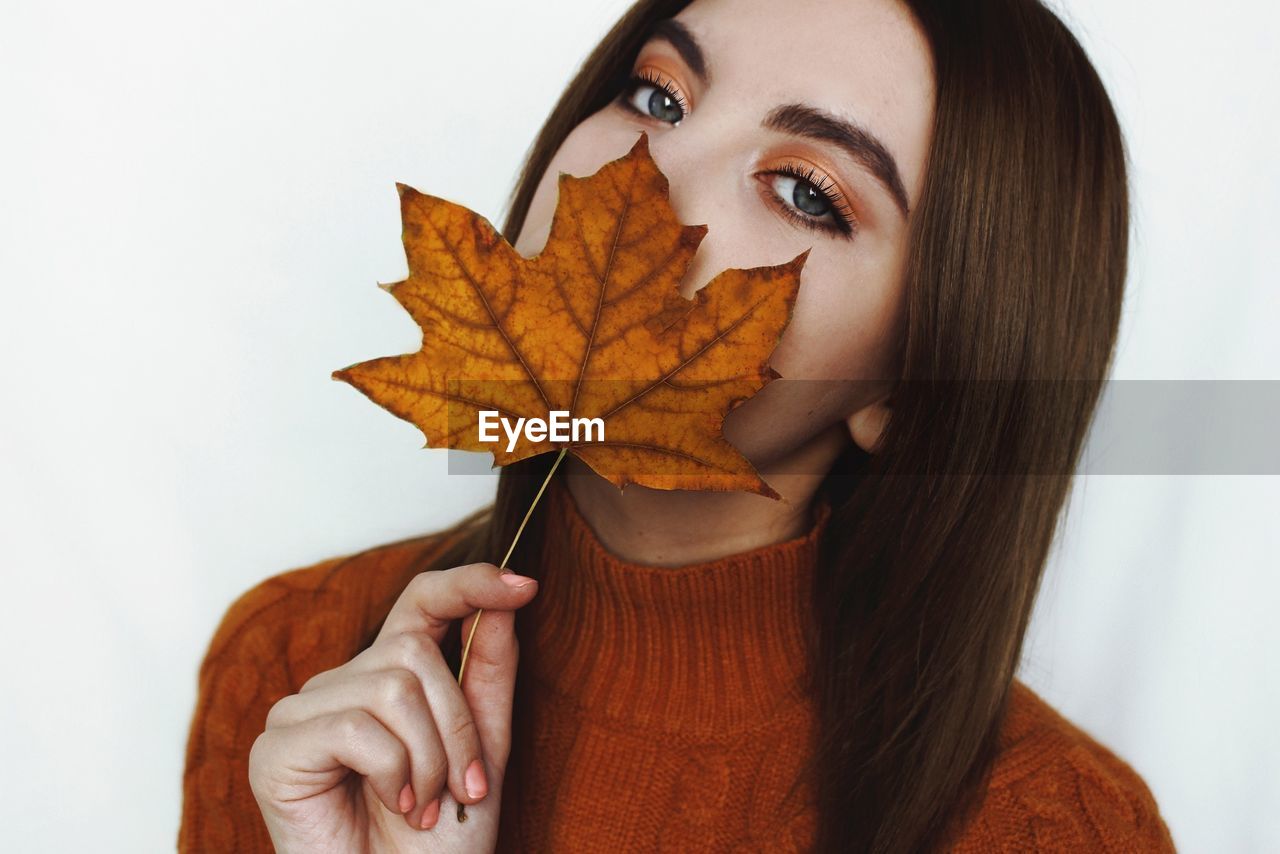 Close-up portrait of young woman holding autumn leaf while standing against white background