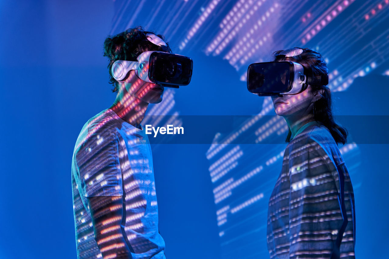 Side view of couple in modern vr goggles while standing on blue background with glowing lights in studio