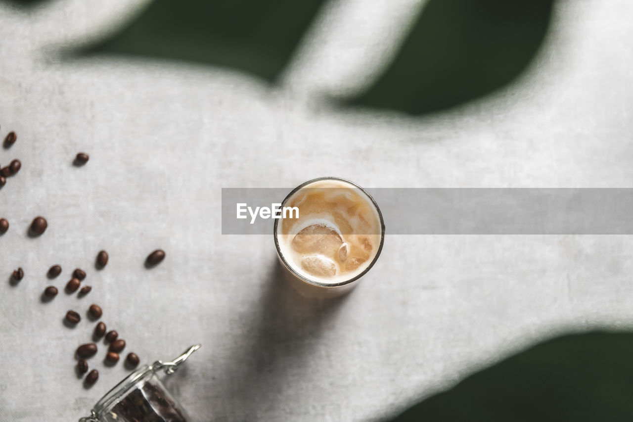 High angle view of iced coffee shot though monsterra on table with roasted cofee beans