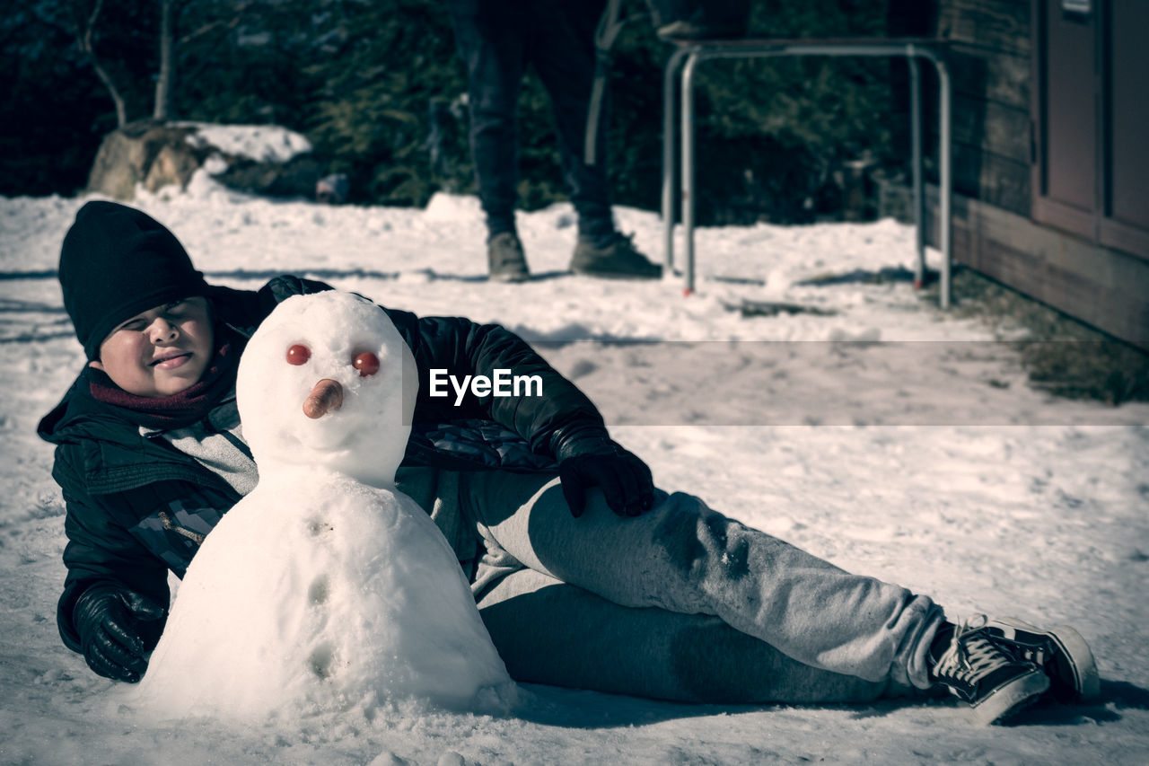 Boy lying in the snow next to self-made snowman with carrot nose and tomato eyes,