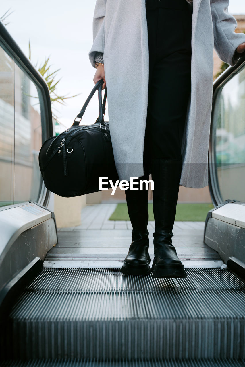 Faceless elegant businesswoman in coat and with bag stepping on escalator in city center
