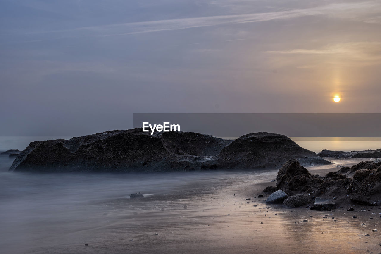 SCENIC VIEW OF ROCKS AT BEACH AGAINST SKY DURING SUNSET