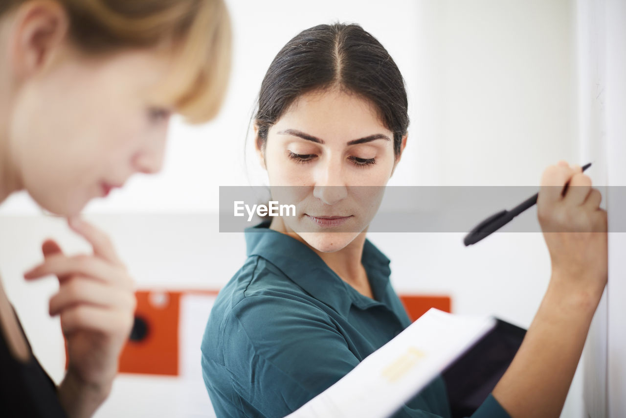 Mid adult businesswoman writing on whiteboard while colleague reading document in office