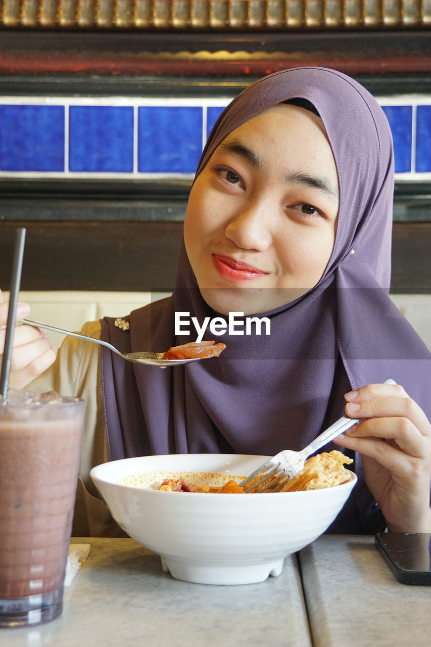 Portrait of young woman wearing hijab while eating food at restaurant