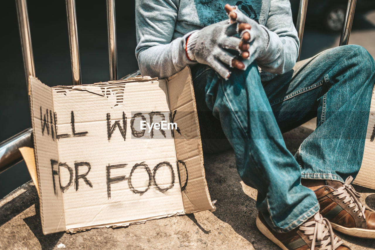 Low section of homeless man with text on cardboard sitting outdoors