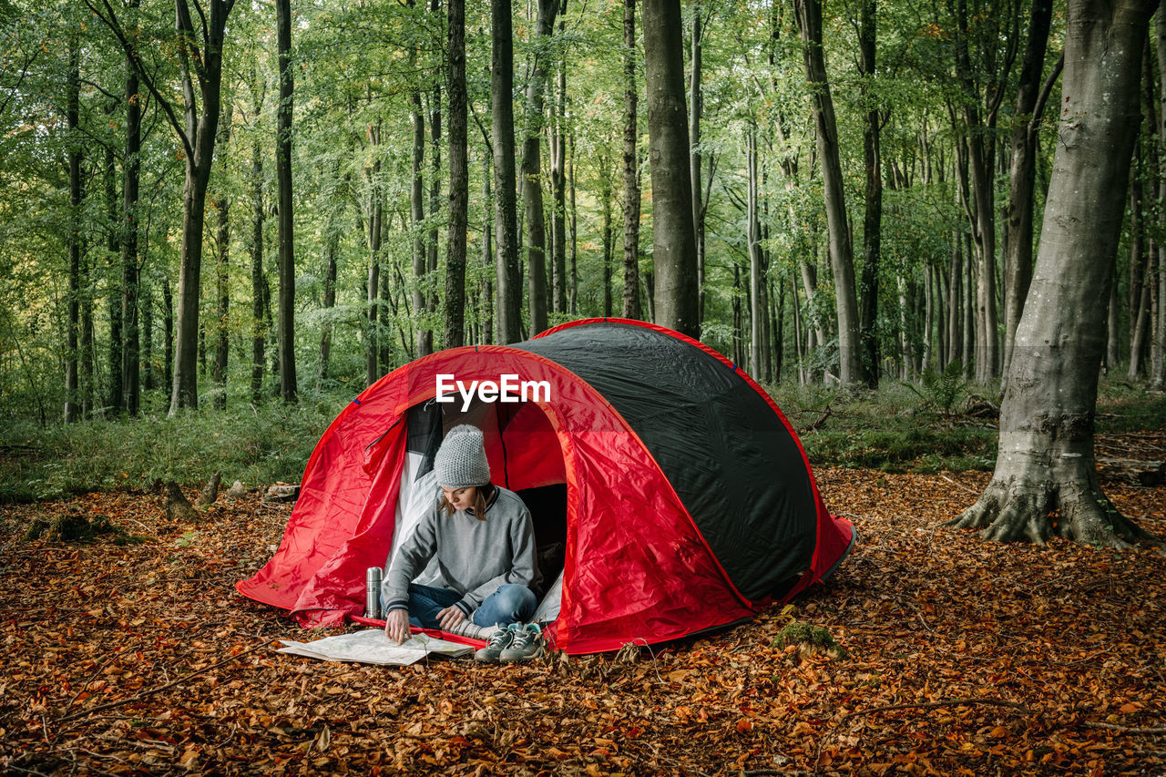 Traveling female sitting near camping tent and navigating with paper map while searching for trail during vacation in forest