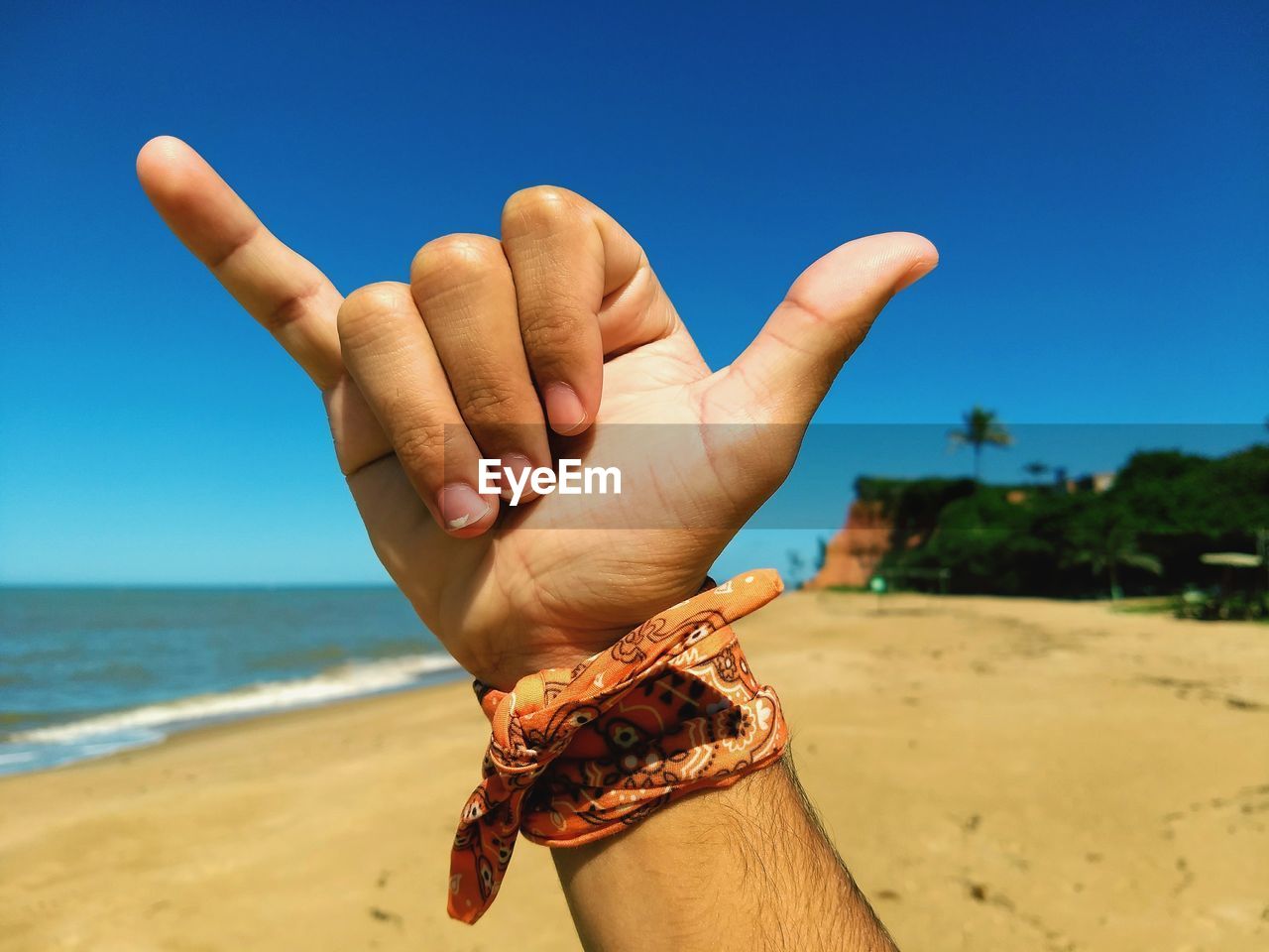 Close-up of man showing shaka sign at beach against blue sky