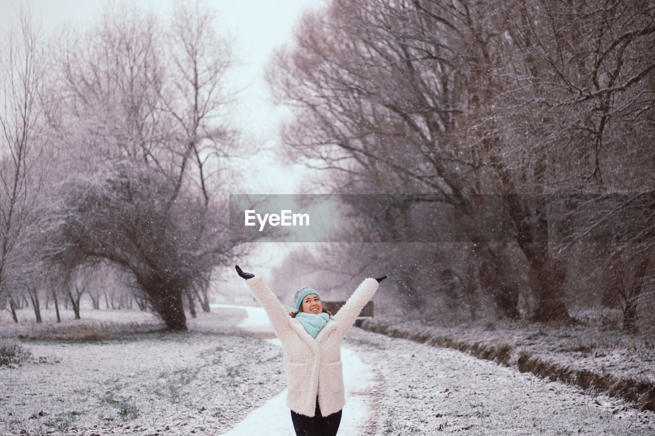 Happy young woman with arms raised standing at park during winter